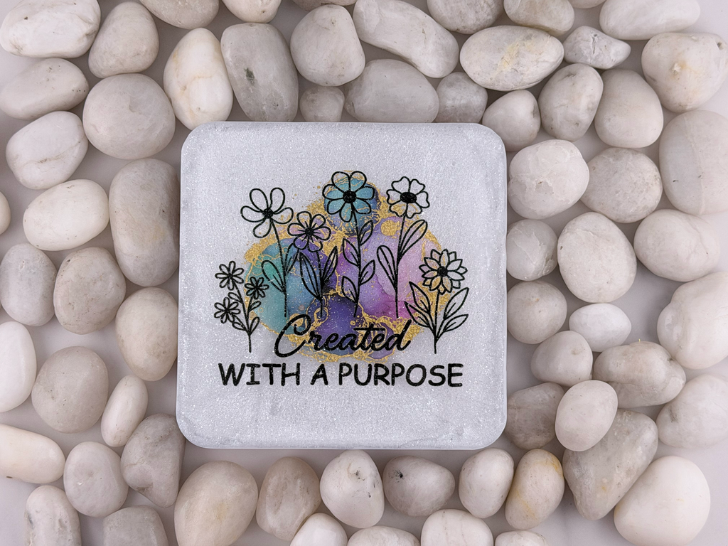 A square stone coaster with rounded corners featuring a black floral illustration and the words 'Created WITH A PURPOSE' over a pastel watercolor background, placed on a bed of smooth white pebbles.