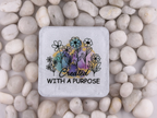 A square stone coaster with rounded corners featuring a black floral illustration and the words 'Created WITH A PURPOSE' over a pastel watercolor background, placed on a bed of smooth white pebbles.