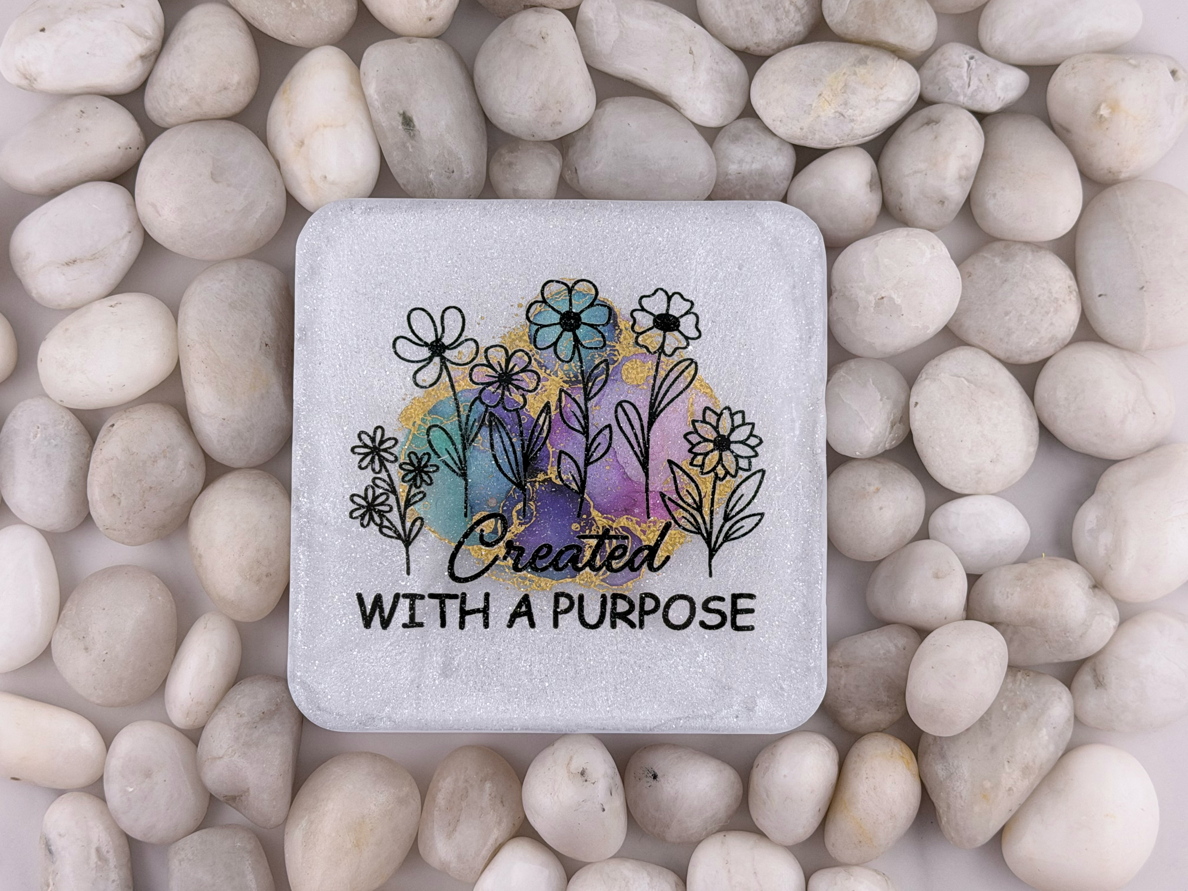 A square stone coaster with rounded corners featuring a black floral illustration and the words 'Created WITH A PURPOSE' over a pastel watercolor background, placed on a bed of smooth white pebbles.