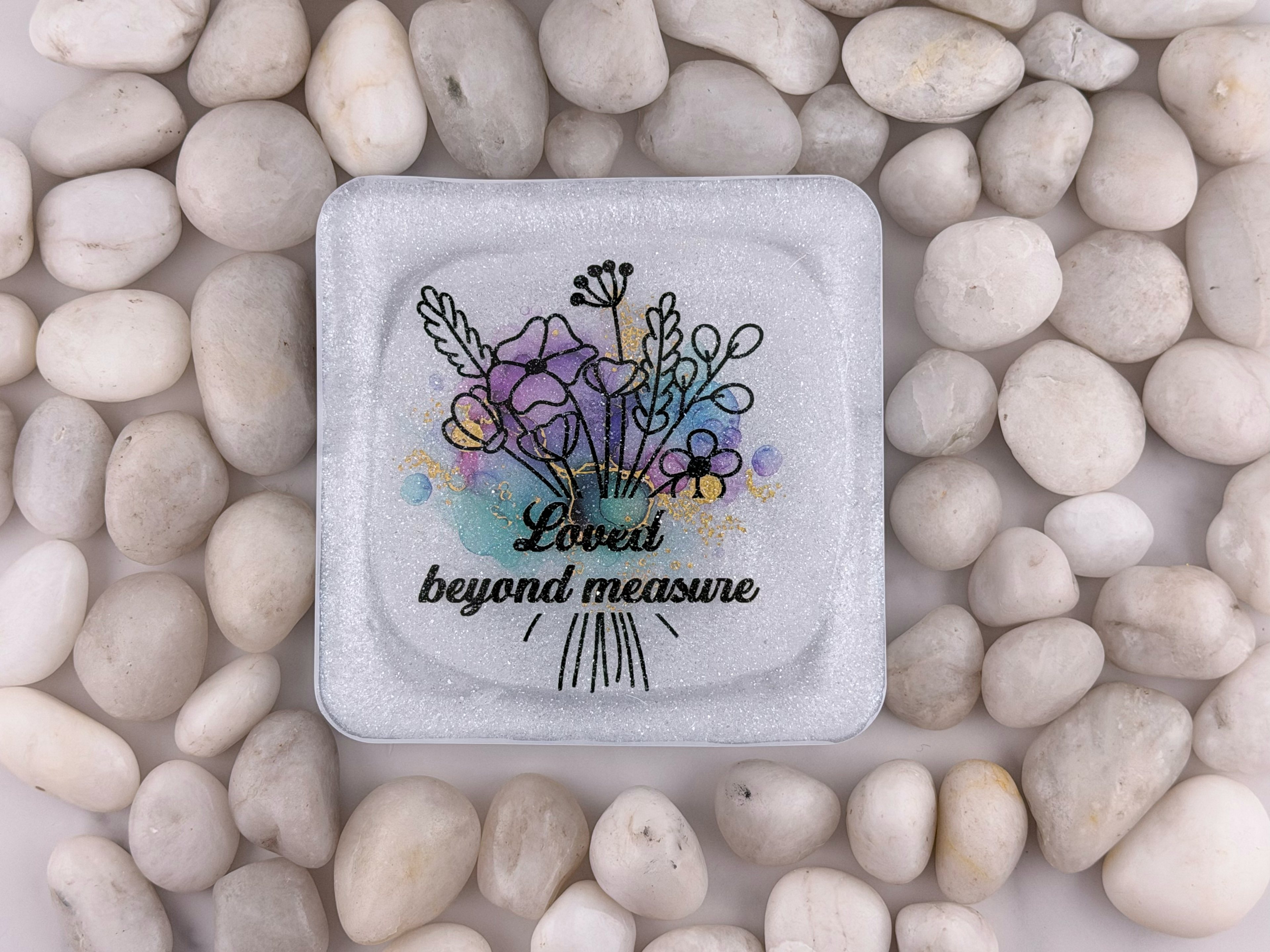 A square glass coaster with rounded corners featuring a floral bouquet illustration and the text 'Loved beyond measure' in black script, surrounded by smooth white pebbles.