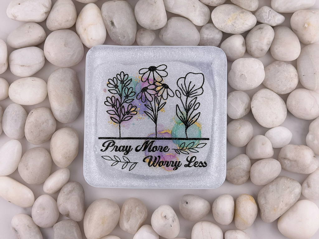 A square glass coaster with rounded corners, featuring a printed design of three stylized flowers and the text 'Pray More Worry Less' in black script. The coaster is placed on a bed of smooth, light-colored river stones.