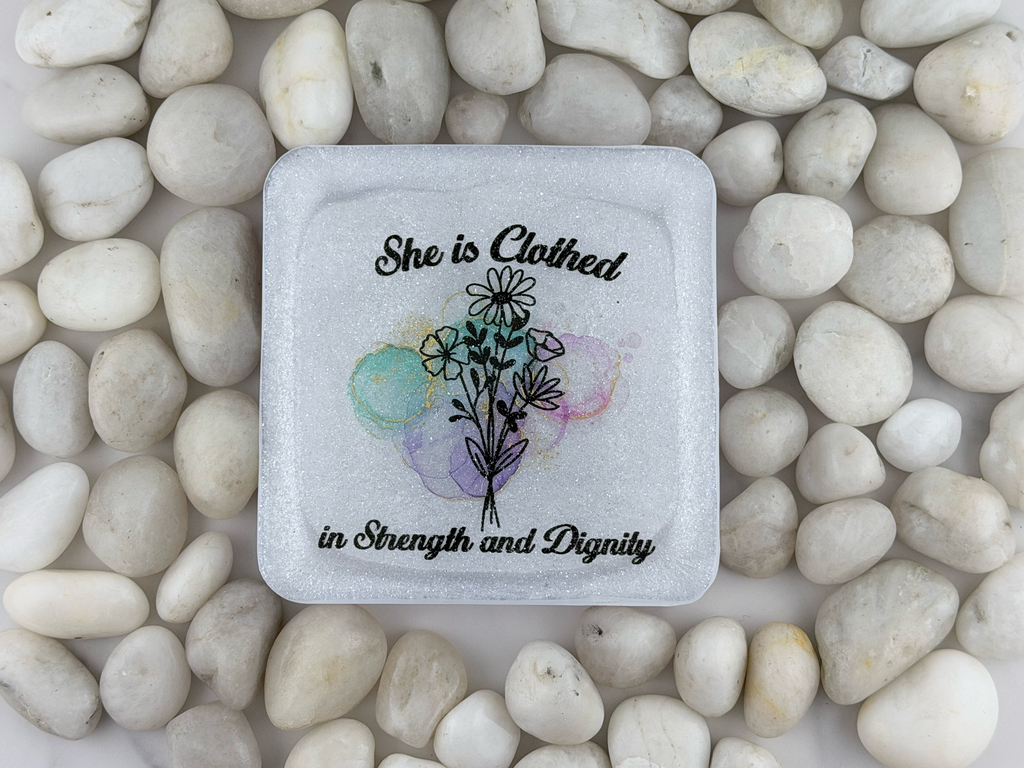 A square white coaster with a printed floral design and the words 'She is clothed in Strength and Dignity,' placed on top of smooth white stones.