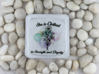 A square white coaster with a printed floral design and the words 'She is clothed in Strength and Dignity,' placed on top of smooth white stones.