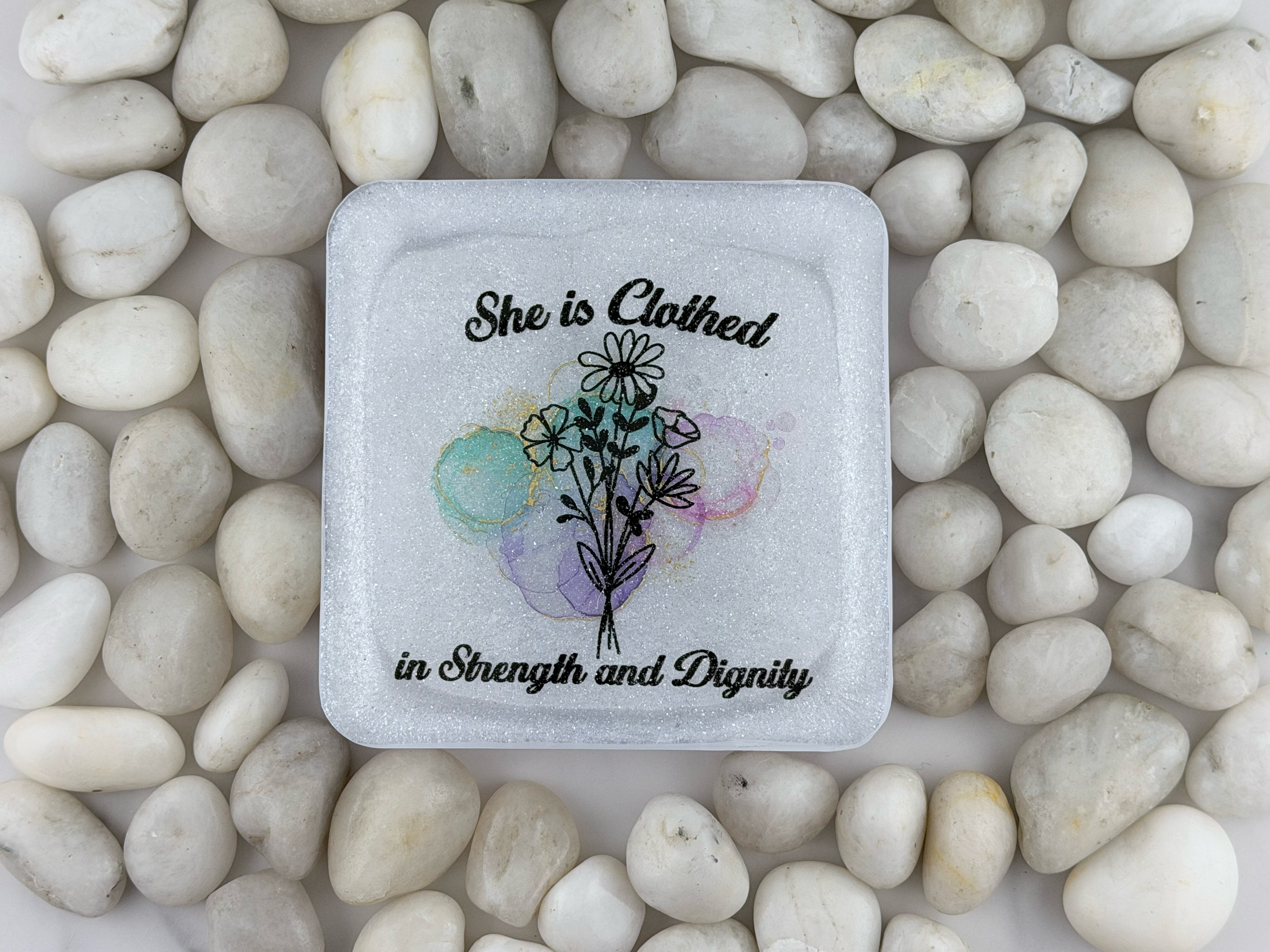 A square white coaster with a printed floral design and the words 'She is clothed in Strength and Dignity,' placed on top of smooth white stones.