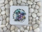A square white coaster with rounded corners, featuring a colorful floral illustration and the text 'God is good all the time' in black script. The coaster is photographed from above on a bed of smooth white pebbles.