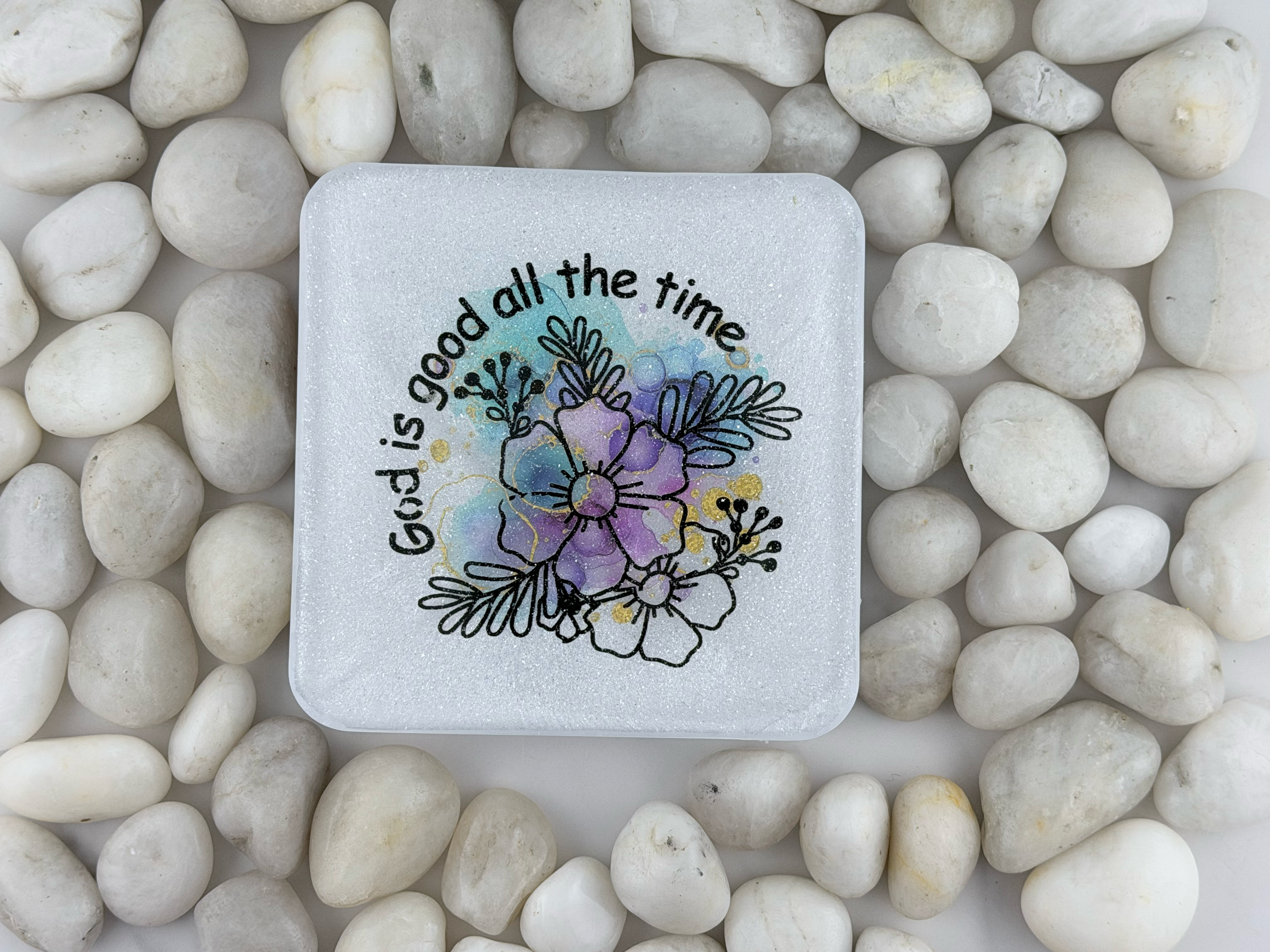 A square white coaster with rounded corners, featuring a colorful floral illustration and the text 'God is good all the time' in black script. The coaster is photographed from above on a bed of smooth white pebbles.