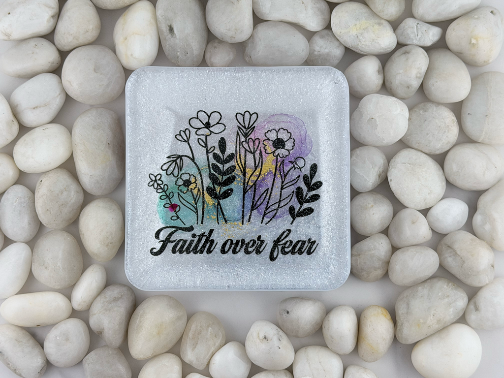 A square frosted coaster with rounded corners, featuring a floral illustration and the words 'Faith over fear' in black script. The plaque is placed on a bed of smooth white pebbles.