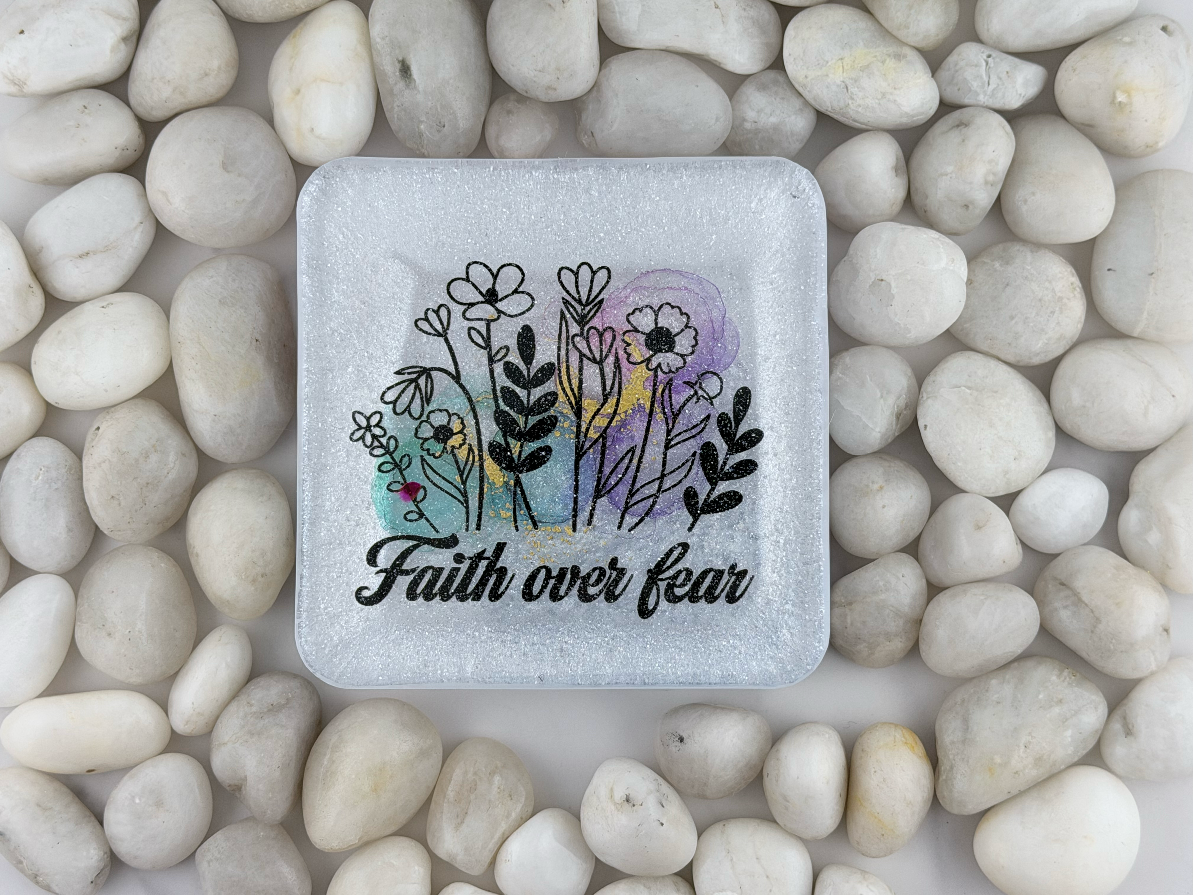 A square frosted coaster with rounded corners, featuring a floral illustration and the words 'Faith over fear' in black script. The plaque is placed on a bed of smooth white pebbles.