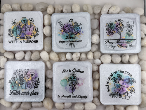 Six square glass coasters with rounded corners arranged in two rows on a bed of small white pebbles. Each coaster features a different inspirational Bible verse in black script and floral illustrations in purple, yellow, blue, and green watercolor styles.