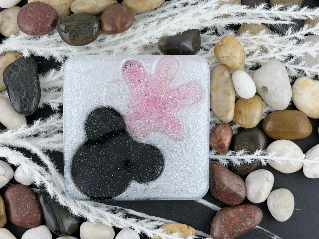 A square clear resin coaster with a frosted white surface, featuring a black abstract organic shape and a pink starfish-like shape. The plaque is photographed from above on a dark background surrounded by smooth multicolored stones and white dried seaweed.