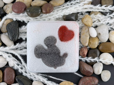 A square white decorative coaster featuring a dark gray Mickey Mouse silhouette and a red heart, surrounded by smooth multicolored stones and white faux spiderwebs on a black surface.