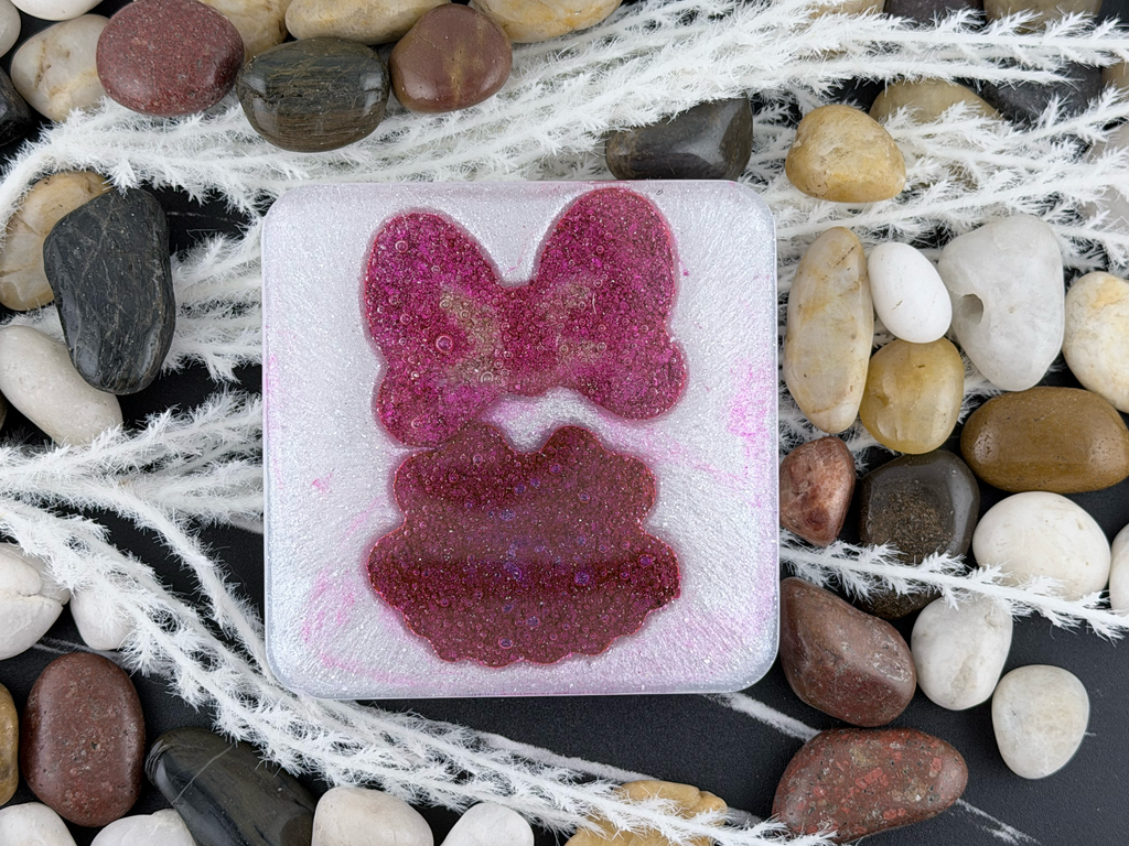 A square translucent resin coaster featuring Minnie Mouse's pink bow and dress, surrounded by smooth river stones and white decorative fiber on a dark surface.