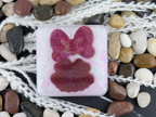 A square translucent resin coaster featuring Minnie Mouse's pink bow and dress, surrounded by smooth river stones and white decorative fiber on a dark surface.