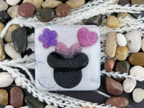 A square handmade resin or glass tile featuring a Minnie Mouse silhouette with a purple flower and pink heart on the head, surrounded by decorative white nettle and smooth river stones.