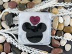 A square coaster featuring a black Mickey Mouse silhouette and a glittery pink heart on a frosted white background, styled with white netting and smooth stones.