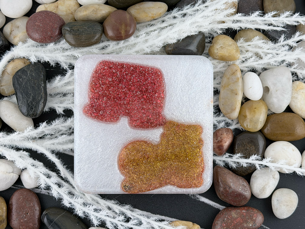 A square white resin coaster featuring Mickey Mouse's shorts and one of his shoes displayed on a bed of smooth river stones and white decorative grass.