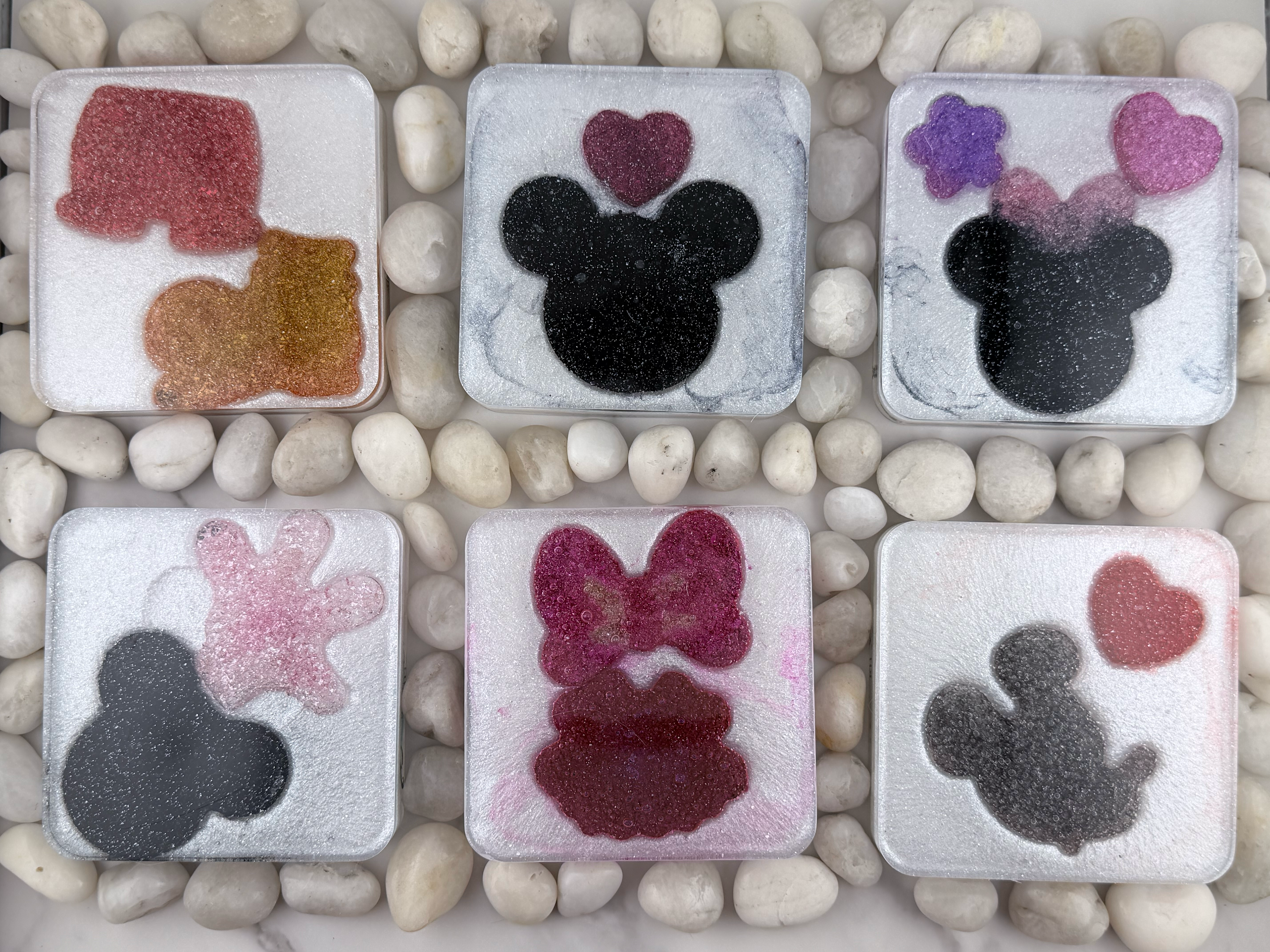 Six square translucent resin tiles arranged in two rows on a bed of small white stones. Each tile features a different Minnie Mouse-inspired design, including a mouse head silhouette, hearts, bows, and glittery accents in pink, purple, black, and red.