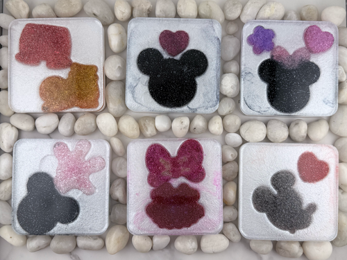 Six square translucent resin tiles arranged in two rows on a bed of small white stones. Each tile features a different Minnie Mouse-inspired design, including a mouse head silhouette, hearts, bows, and glittery accents in pink, purple, black, and red.
